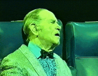 an elderly man in a sweater and bow tie is sitting in a chair with his mouth open