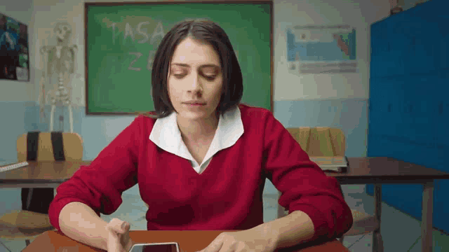 a woman in a red sweater sits at a desk in front of a blackboard that says pasa