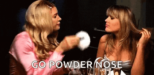 two women are sitting at a table talking to each other while one of them is holding a powder nose .