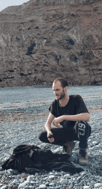 a man squatting on a rocky beach wearing a black shirt with the number 19 on his pants