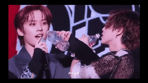 a man is drinking water from a bottle while another man holds a glass .
