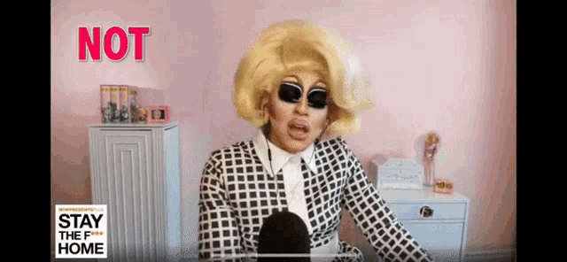 a drag queen is talking into a microphone while wearing sunglasses and a plaid suit with the words " not " behind her