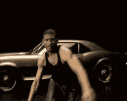 a man in a black tank top is dancing in front of a silver car .
