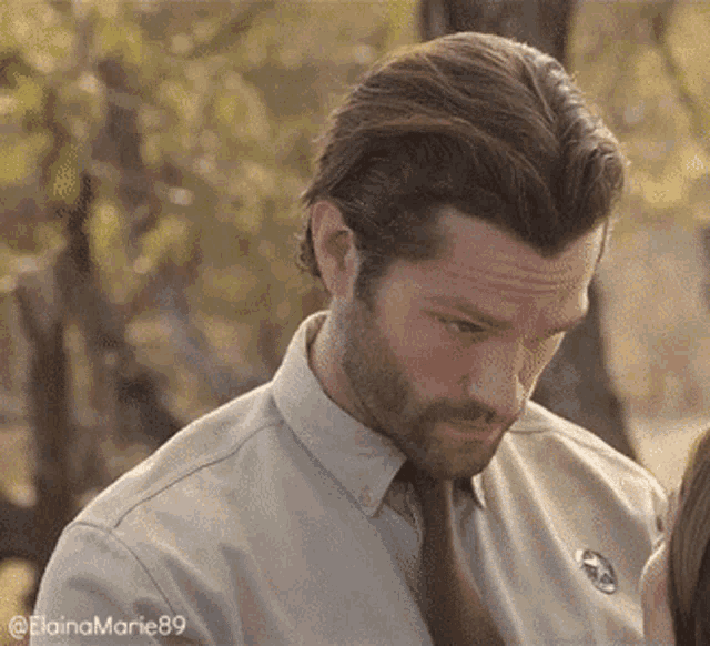 a man with a beard is wearing a shirt and tie and looking down .