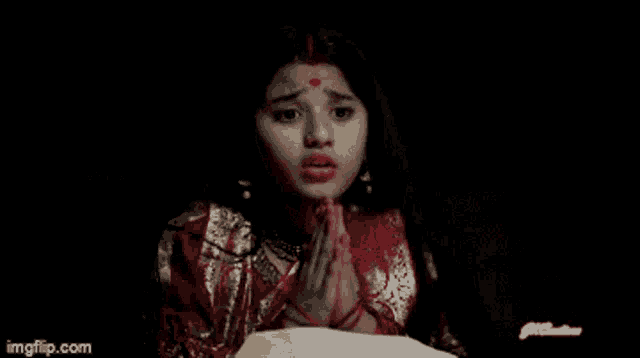 a girl in a red and gold dress is praying in a dark room