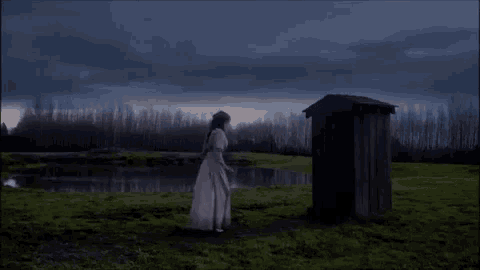 a woman in a white dress stands in front of a wooden outhouse in a field
