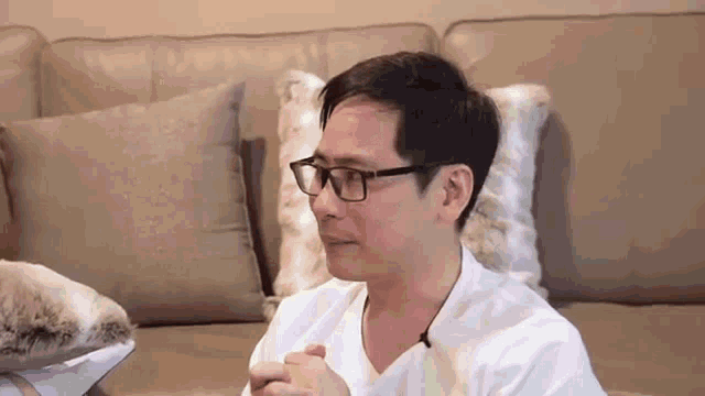 a man wearing glasses is sitting on a couch with his hands folded in front of him .