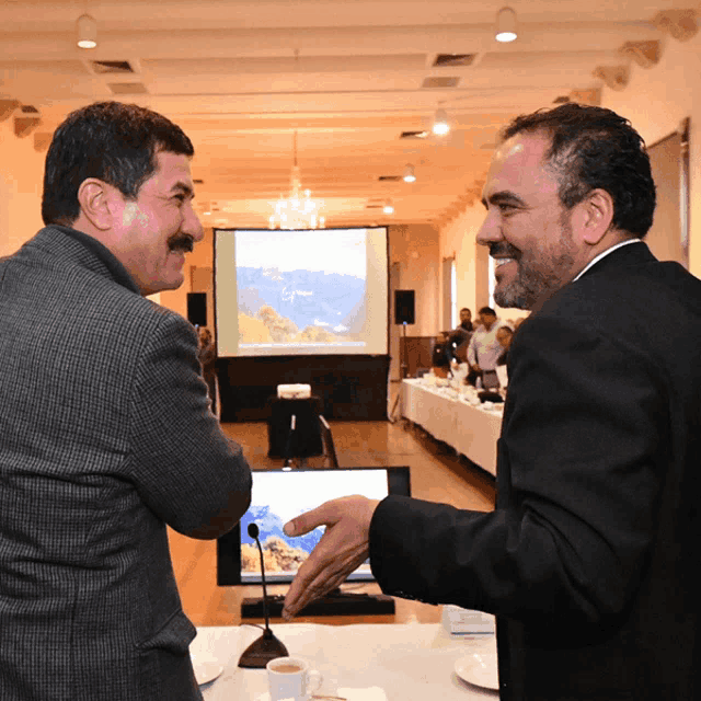 two men shake hands in front of a large screen