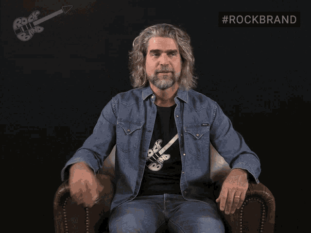 a man with long hair and a beard is sitting in a chair in front of a sign that says rockbrand