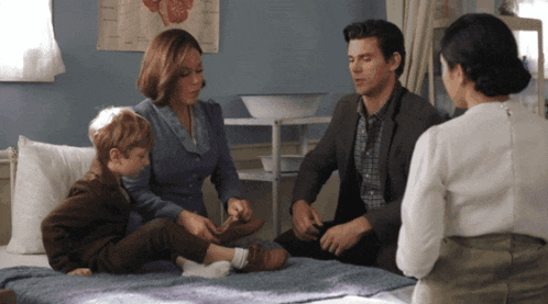 a family sits on a bed talking to a doctor while a poster of a heart hangs on the wall behind them