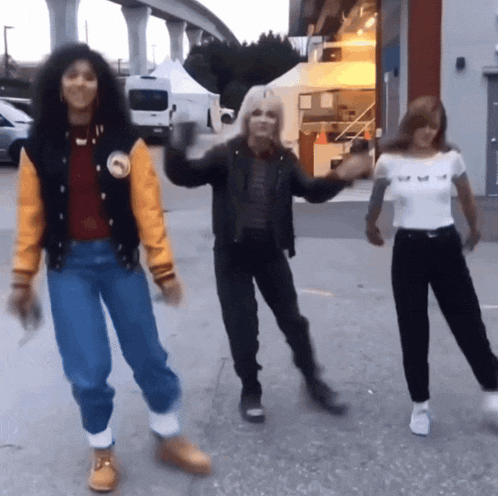 three women are dancing in a parking lot and one has a yellow jacket