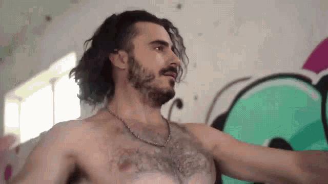 a shirtless man with long hair and a beard stands in front of a wall with graffiti on it