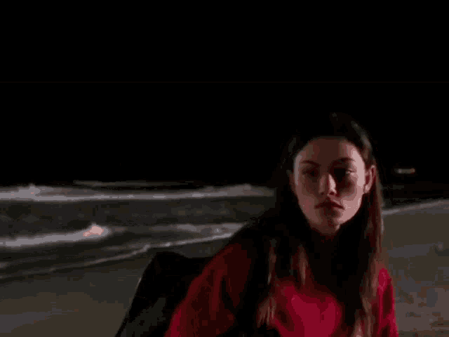 a woman in a red sweater is sitting on a beach at night looking at the ocean .