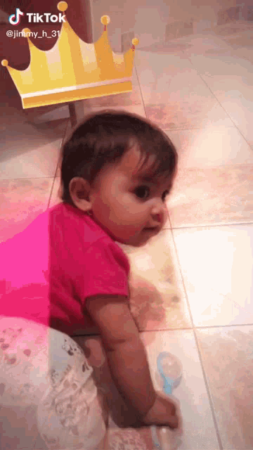 a baby in a pink shirt is crawling on the floor next to a crown that says tiktok on it