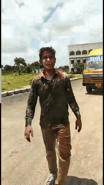 a man in a dirty shirt is walking down a street in front of a school bus that says shah