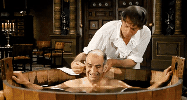a bald man is being shaved in a wooden tub by another man