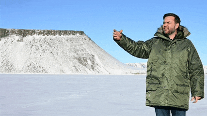 a man in a green parka stands in front of a mountain