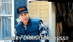 a man wearing a hat and a vest is standing in front of a house and says merry christmas russ .