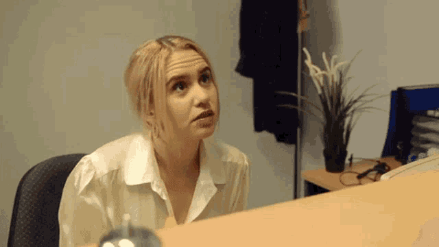 a woman in a white shirt is sitting at a desk with a plant in the background