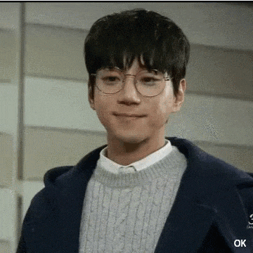 a young man wearing glasses and a sweater is smiling while standing in front of a wall .