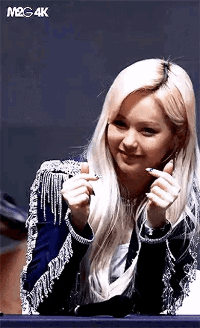 a woman with blonde hair is making a heart shape with her hands while sitting at a table .