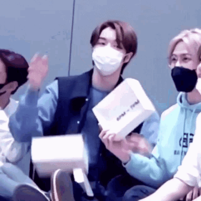a man wearing a mask is holding a box in his hand while sitting in a room with other people .