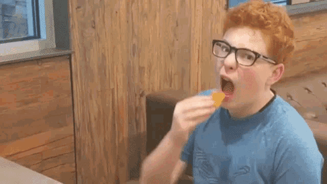 a young man wearing glasses and a blue shirt is eating something with his mouth open .