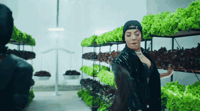 a woman in a black hat is standing in front of a row of green plants