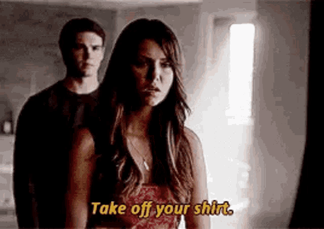 a man and a woman are standing next to each other in a dark room and the woman is saying `` take off your shirt . ''