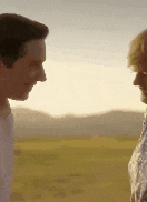 a man and a woman are looking at each other in a field with mountains in the background .