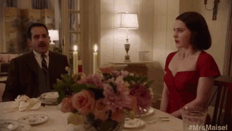 a woman in a red dress sits at a table with a man in a suit behind her