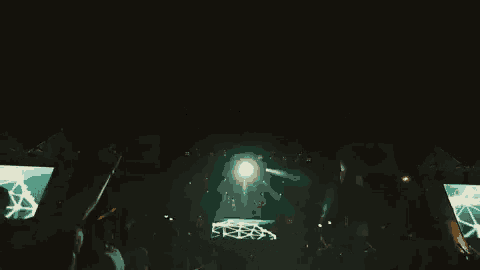 a crowd of people are standing in front of a stage at a concert with lights coming out of it .