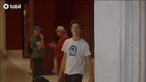 a man in a white t-shirt is walking down a hallway with a tabii logo in the background