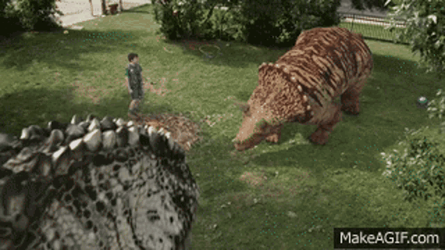 a boy is standing next to a dinosaur in a park and the website makeagif.com is visible in the corner