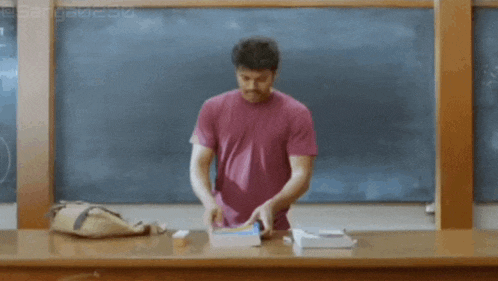 a man in a pink shirt is reading a book in front of a blackboard in a classroom .