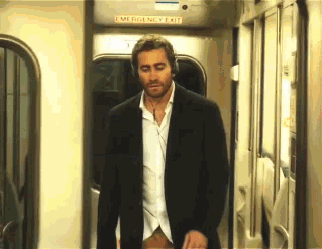 a man wearing headphones is walking on a train with an emergency exit sign above him