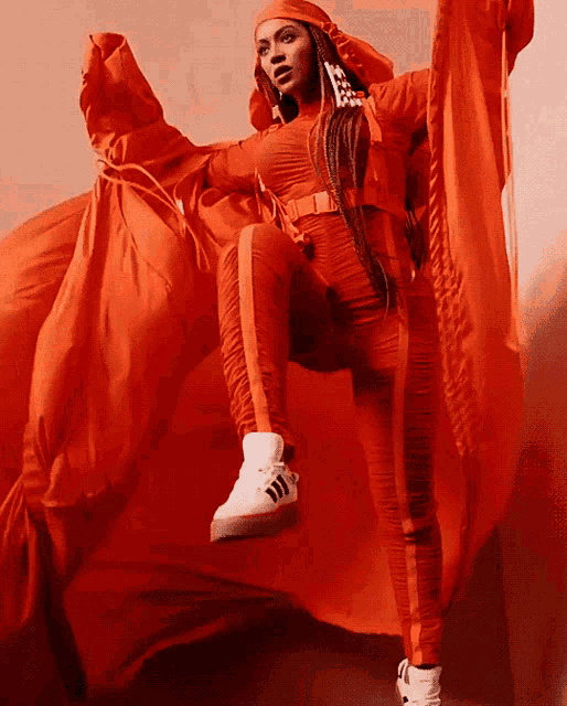 a woman wearing an orange outfit and white adidas shoes stands in front of an orange cloth