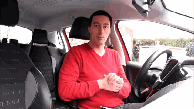 a man in a red sweater sits in a car with his hands folded