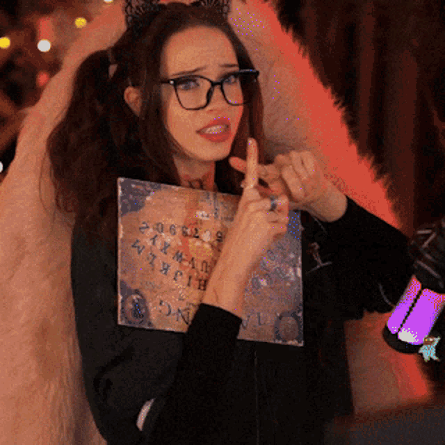 a woman wearing glasses and a cat ear headband is holding a piece of paper that says ouija on it