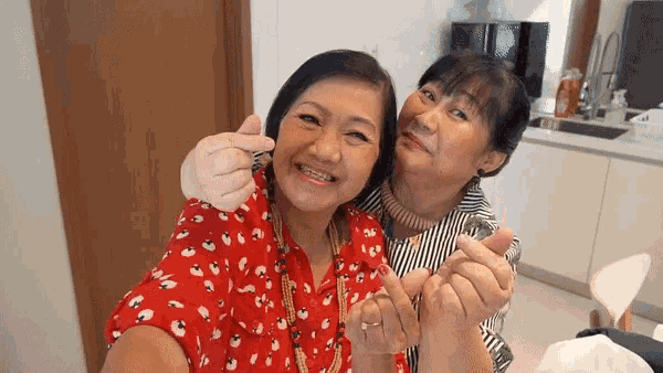 two older women are hugging each other in a kitchen and making a heart with their hands .