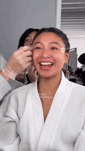 a woman in a bathrobe is smiling while getting her makeup done .