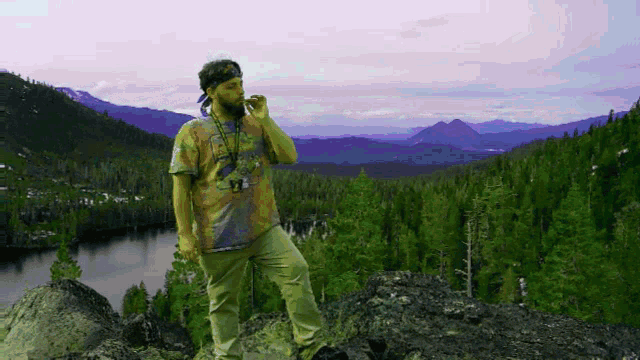 a man in a tie dye shirt is standing on top of a rocky hill overlooking a lake