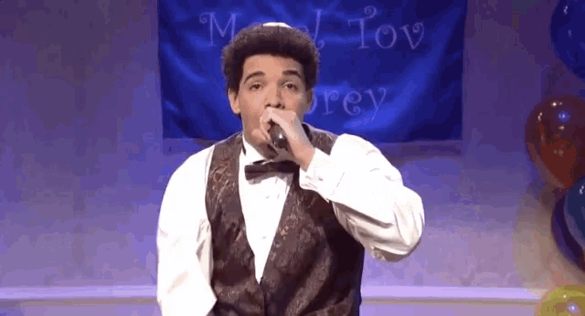 a man in a tuxedo is singing into a microphone in front of a banner that says mat tov rey