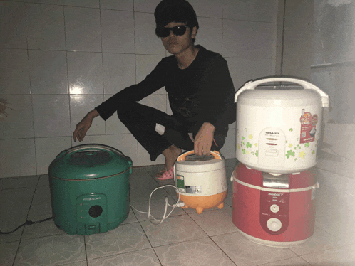 a man squatting next to a stack of rice cookers including one that says sharp
