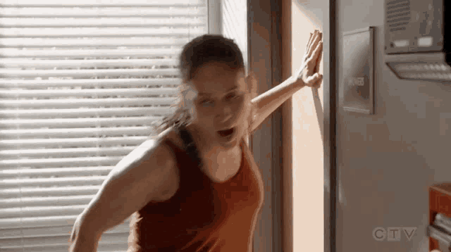 a woman in a red tank top is standing in front of a window with blinds on it .