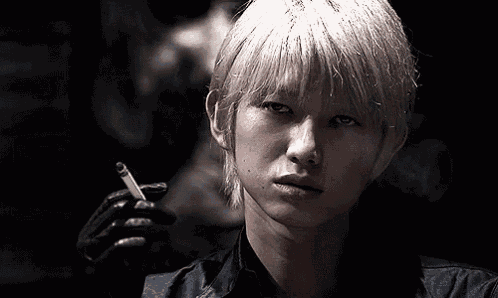 a young man with blonde hair is smoking a cigarette in a dark room .