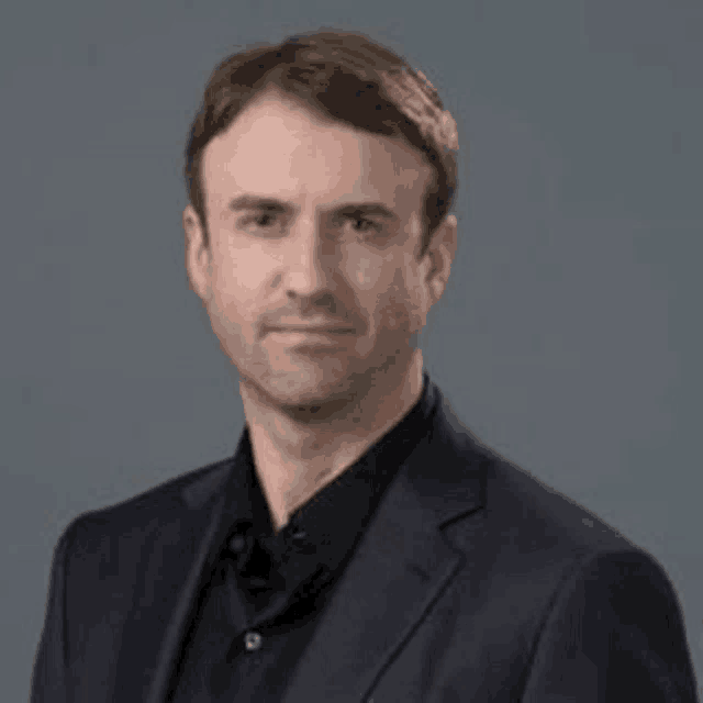 a man in a suit and black shirt is standing in front of a gray wall .