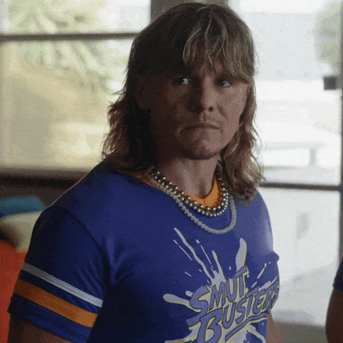 a man with a mullet wears a shirt that says smut busters