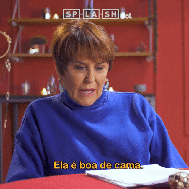 a woman in a blue sweater is sitting at a table and says ela é boa de cama