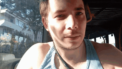 a man wearing a blue tank top and a necklace looks at the camera